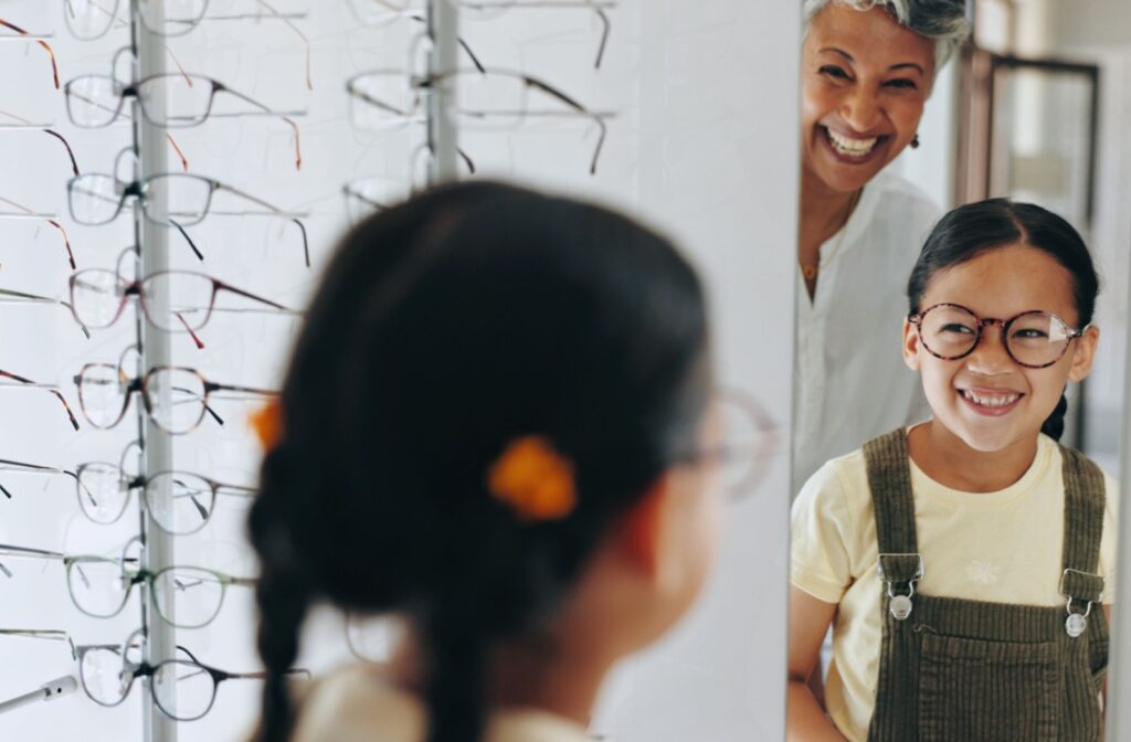 An adult and child smiling in a mirror in an optometry clinic while browsing and trying on new glasses