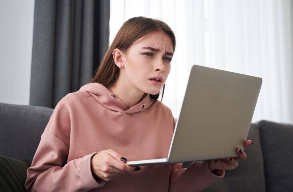 An adult sits on a couch, leaning forward and squinting while holding a laptop close to her face.