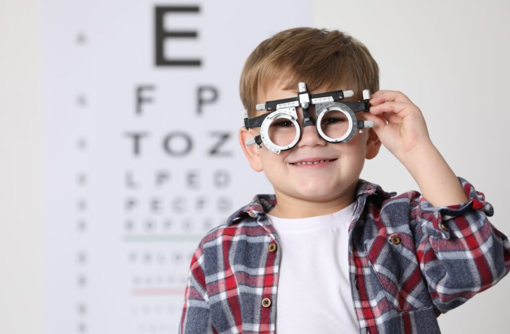 A smiling child wears a trial lens frame during an eye exam, standing in front of a blurred vision chart.