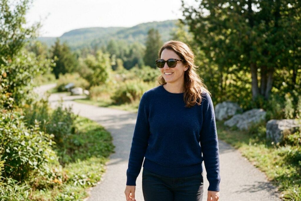 Person in sunglasses and navy sweater walking on a paved path, smiling, surrounded by greenery with forested hills behind.