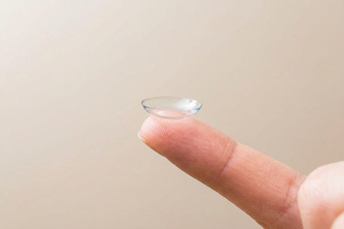 Contact lens balanced on the tip of a fingertip against a plain beige background.